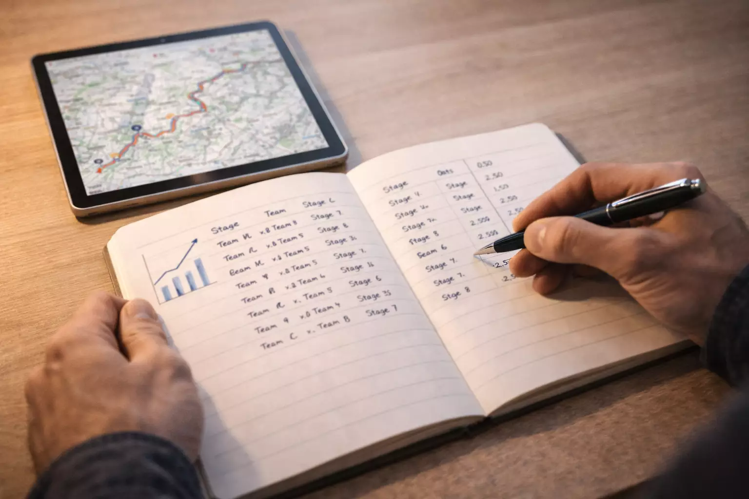 Persona revisando un cuaderno con anotaciones de apuestas de ciclismo junto a una pantalla con etapas de una gran vuelta